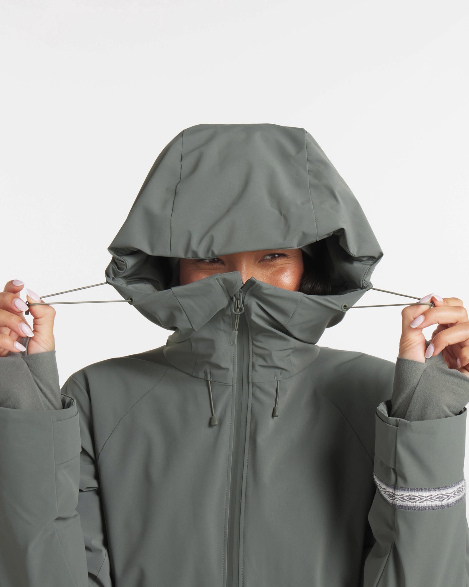 Front close-up of Women’s Apu Parka with hood cinched, high zipped collar, and thumbhole cuffs on light gray background