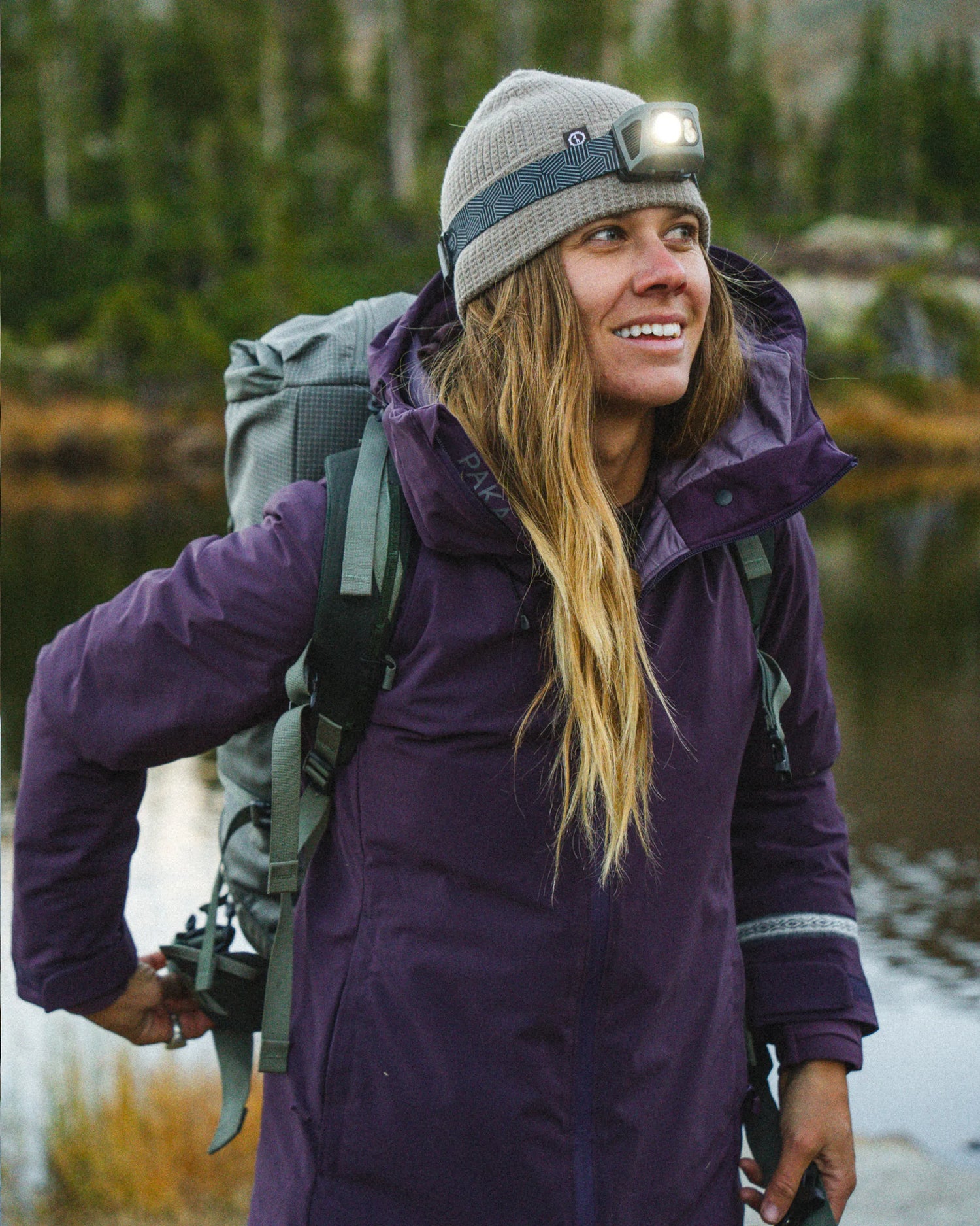 Woman with Backpack and Headlamp in Amethyst / Purple Apu Parka in Front of Lake in Forest – PAKA Apparel