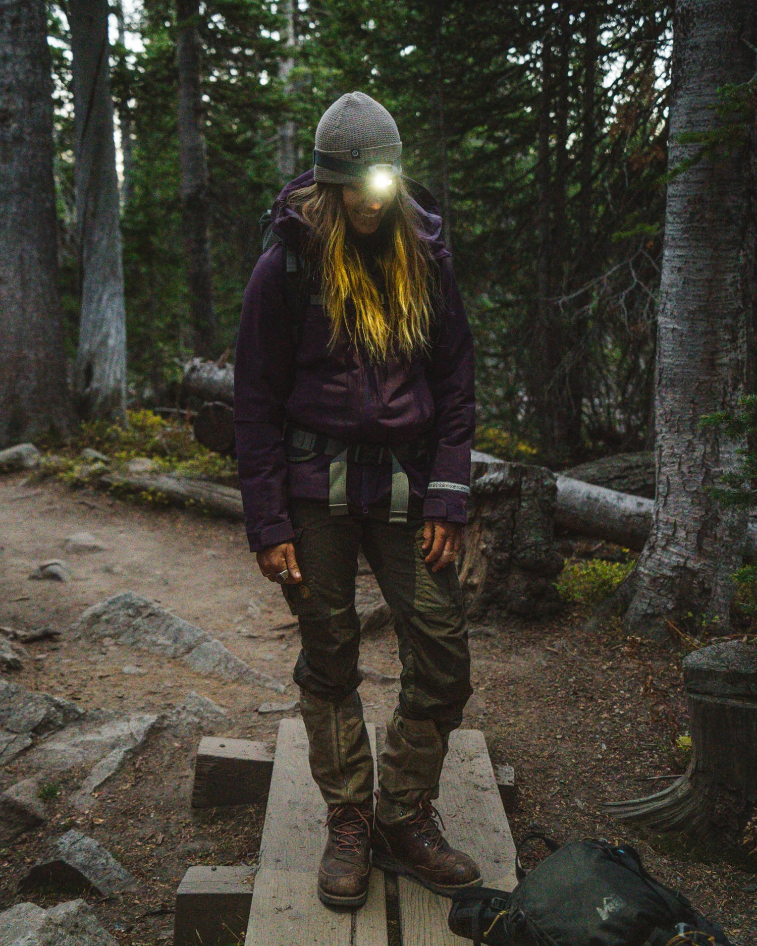 Woman Crossing Small Wood Bridge with Lit Headlamp Wearing Amethyst / Purple Apu Parka – PAKA Apparel