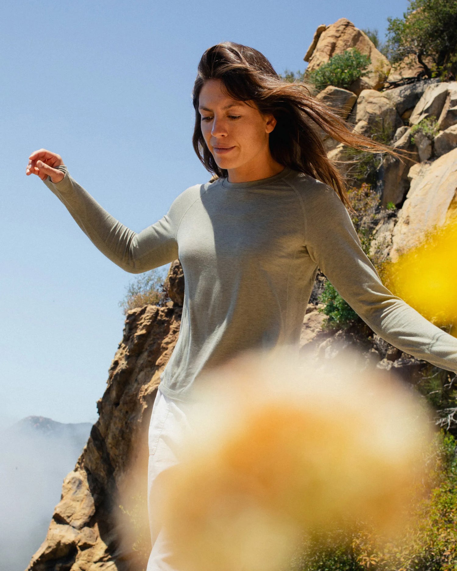 Woman in a light green long-sleeve top and white pants standing on rocky terrain with a clear blue sky.