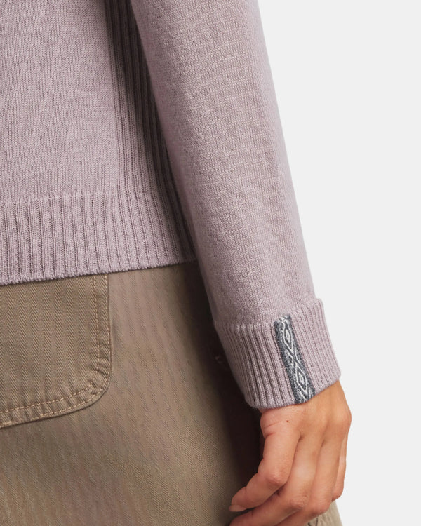 Close-up of a person wearing a light purple sweater with a brand logo on the sleeve, against a neutral background.