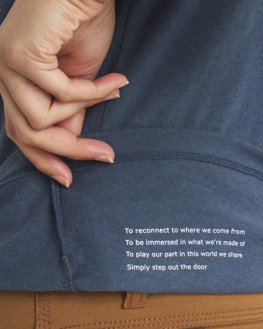 Close-up of Essential V-Neck t-shirt hem showing printed line ‘To reconnect to where we come from’ with hand lifting hem against brown pants