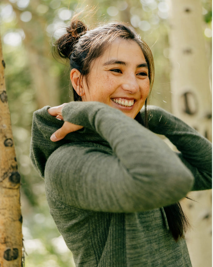 Woman smiling pulls hood on wearing eucalyptus green lightweight alpaca hoodie in a sunlit forest setting