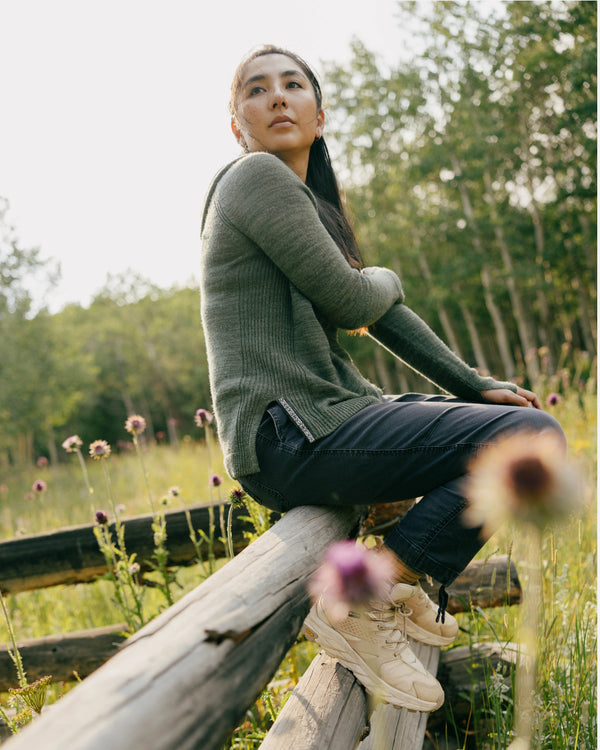 Woman sitting on a wooden fence wearing eucalyptus lightweight alpaca hoodie and dark pants in a sunlit wildflower meadow