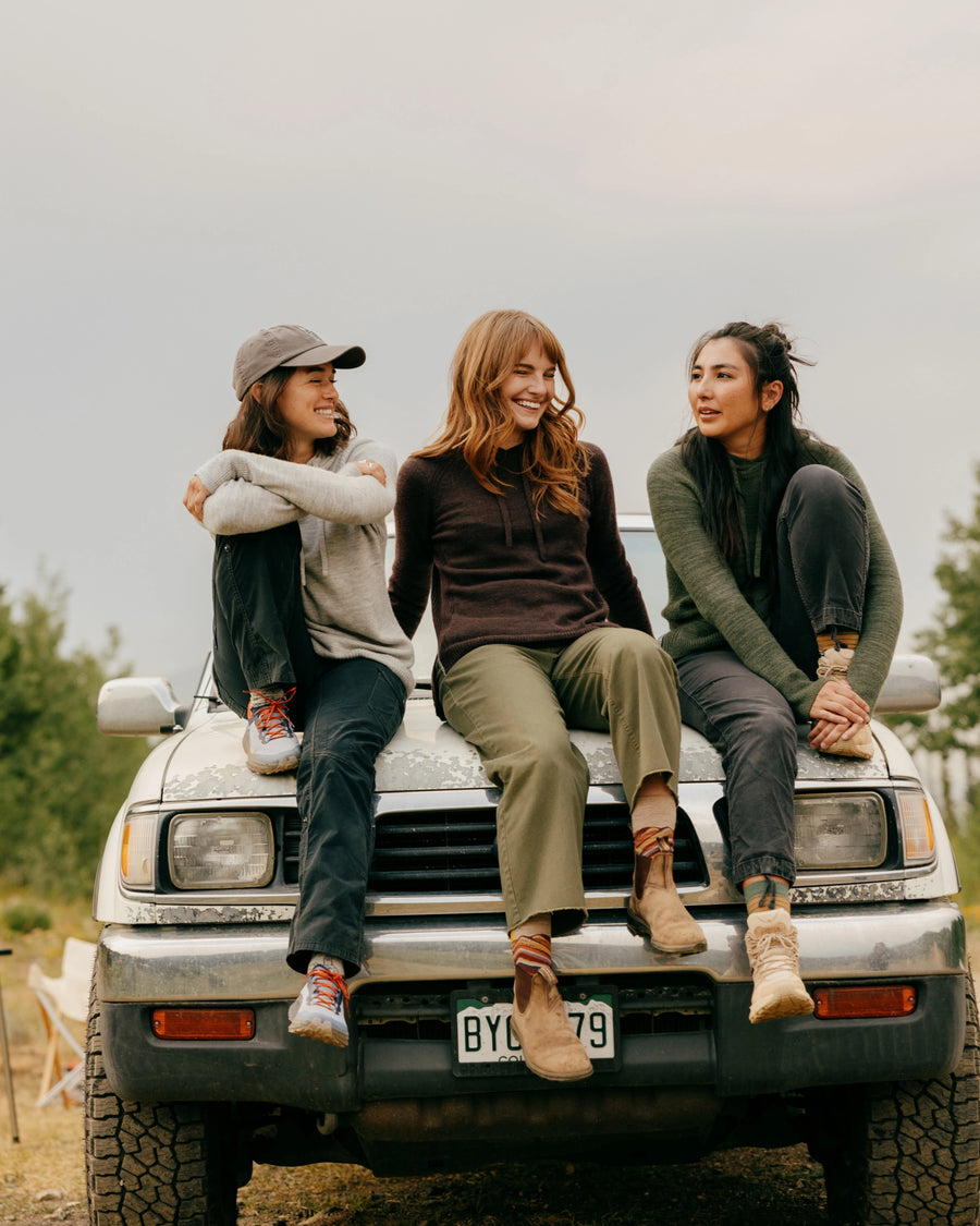 Three women sitting on a pickup truck hood wearing alpaca hoodies in gray, dark brown, and eucalyptus at an outdoor campsite