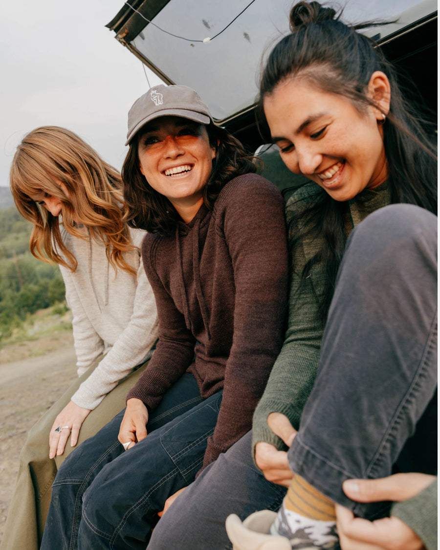 Three women wearing lightweight alpaca hoodies laugh while sitting on a car tailgate in an outdoor travel setting