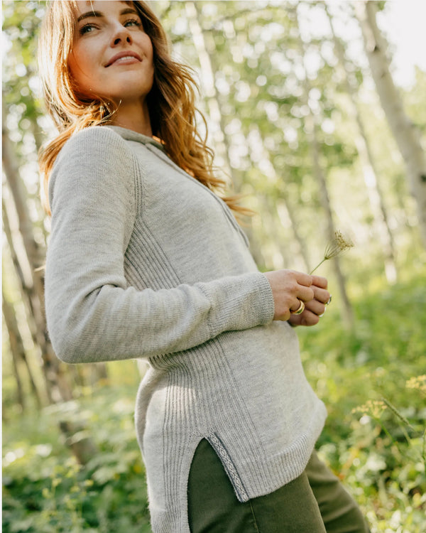Three-quarter side view of women’s alpaca hoodie in light grey showcasing ribbed side panels and relaxed fit on model in a sunlit forest