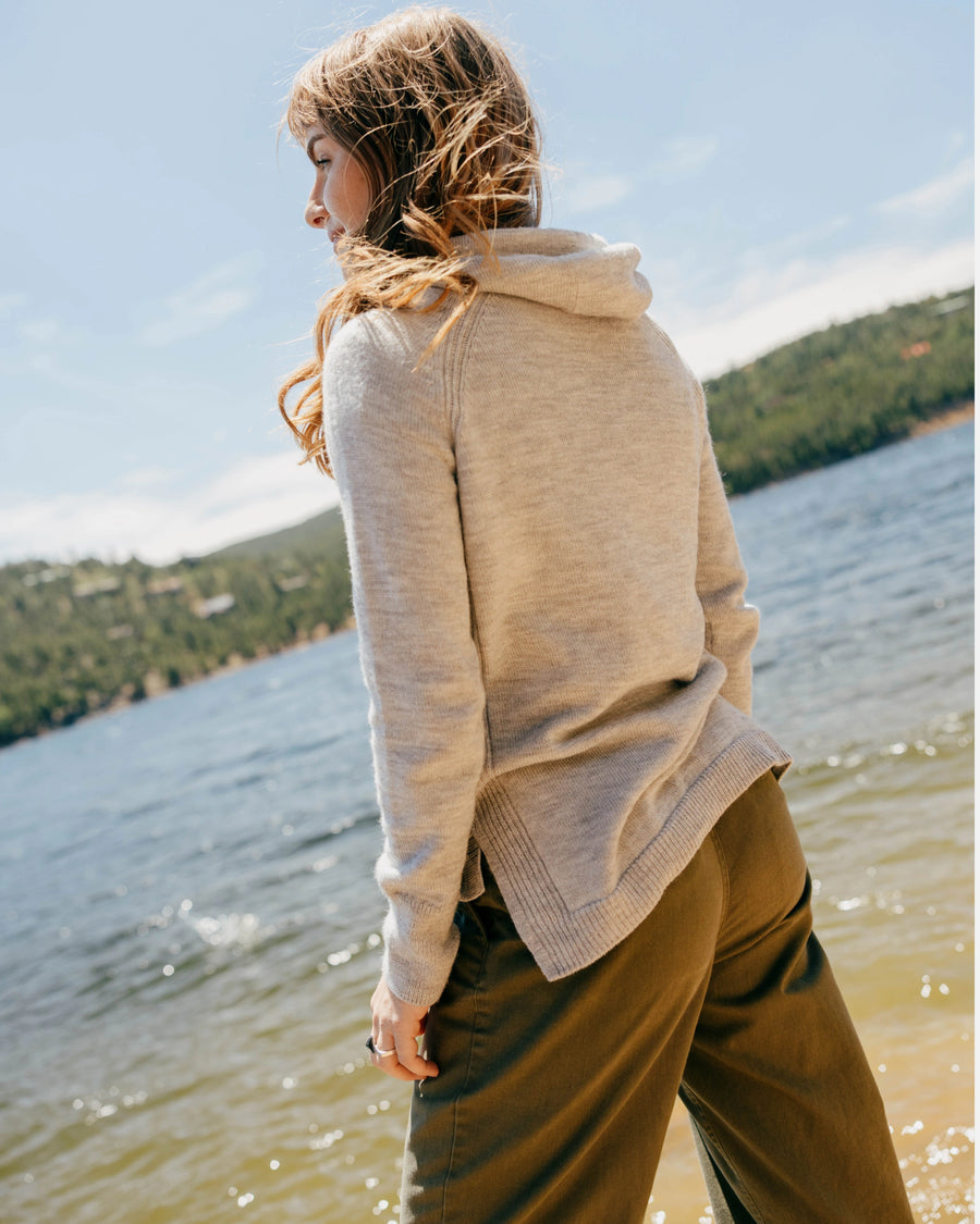 Woman standing at a lakeshore wearing a light grey alpaca hoodie, back view showing relaxed fit and side hem detail