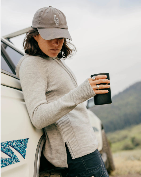 Woman wearing a light grey alpaca hoodie in a relaxed fit leaning against a truck, holding a black travel mug on a hillside