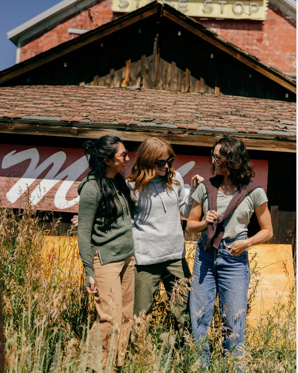 Three women chatting outdoors, center woman wearing a light grey alpaca hoodie in a relaxed fit in front of a rustic barn