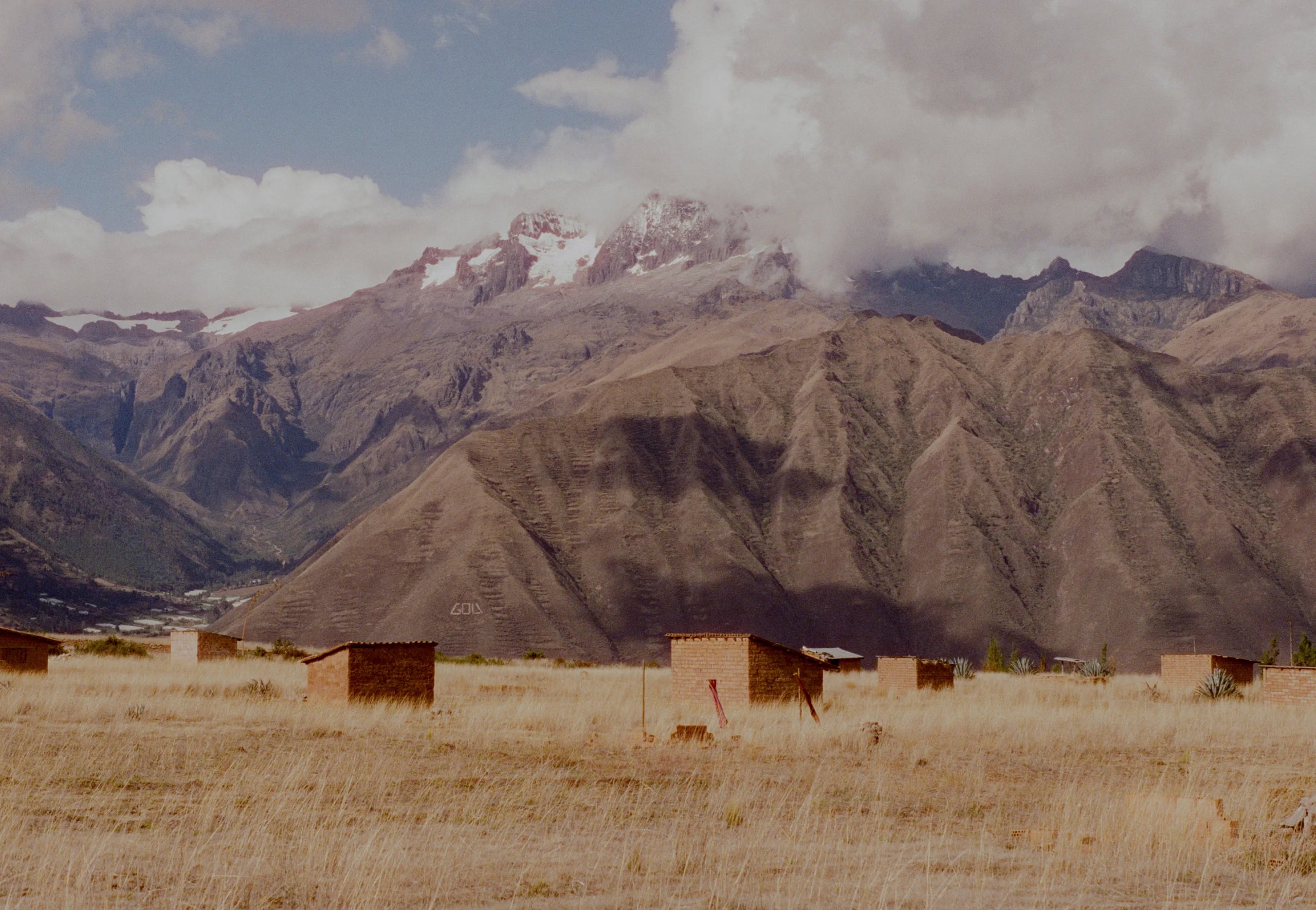 Two small adobe huts sit on a grassy plain beneath towering Andes mountains and a cloudy sky