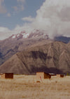 Two small adobe huts nestled in a dry Andean valley with towering snow capped mountains and a dramatic cloudy sky