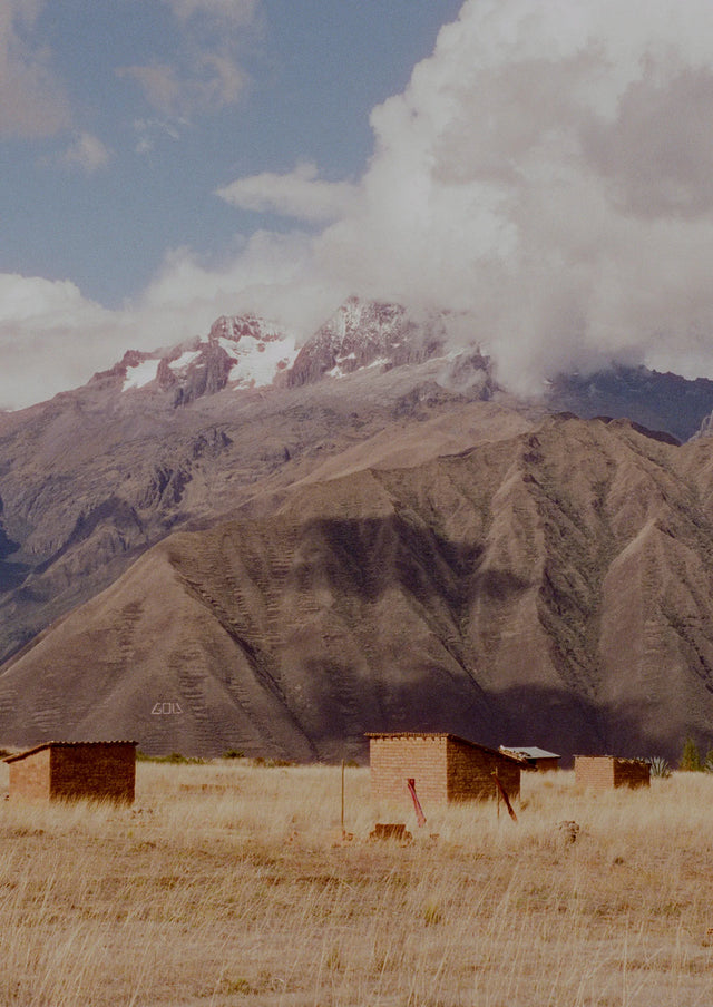Two small adobe huts nestled in a dry Andean valley with towering snow capped mountains and a dramatic cloudy sky