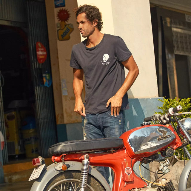 Man standing next to a red Honda motorcycle wearing a black T-shirt on a sunlit street in front of a shop