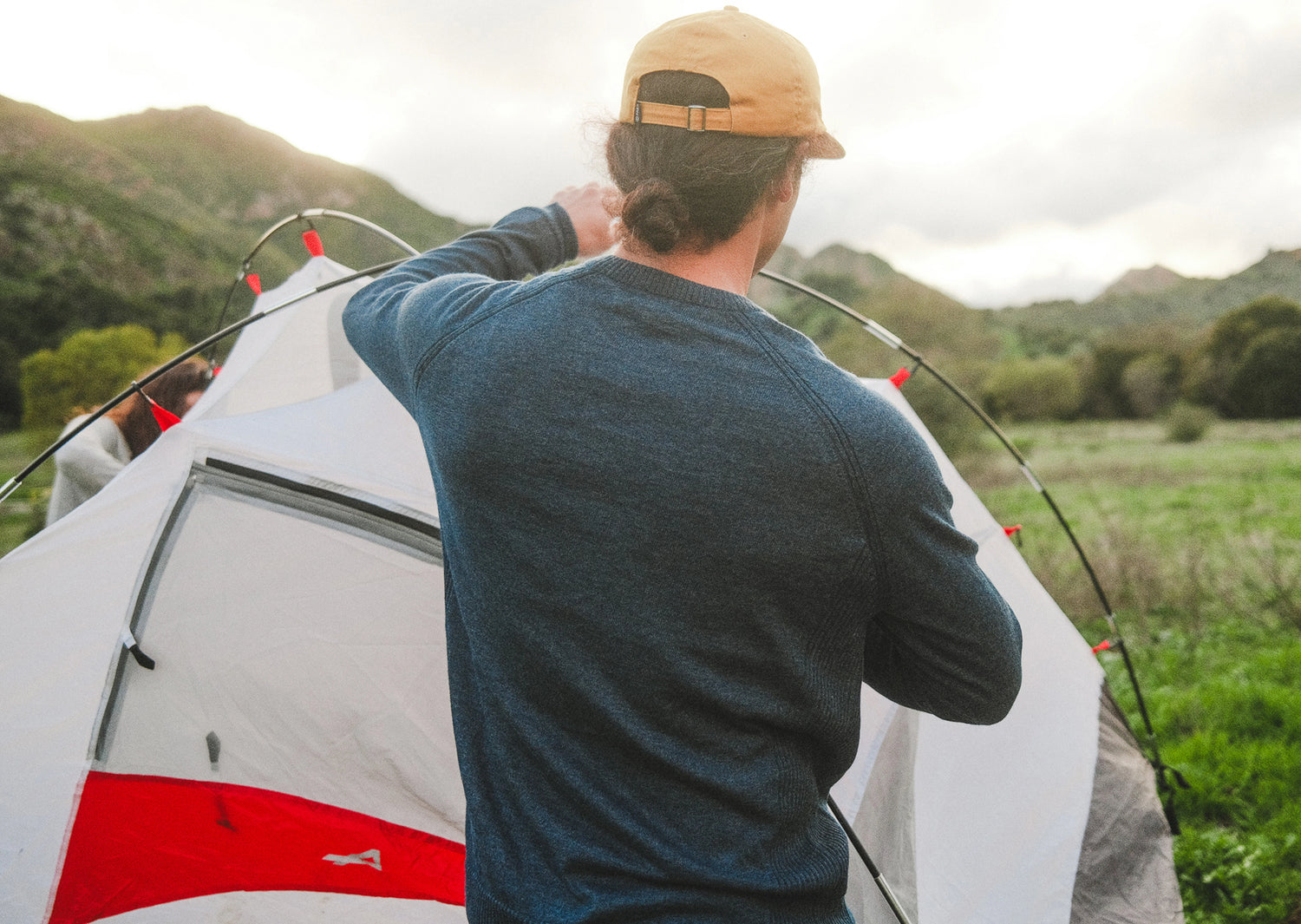 Man wearing a navy ultralight crewneck sweater setting up a tent in a grassy mountain campsite