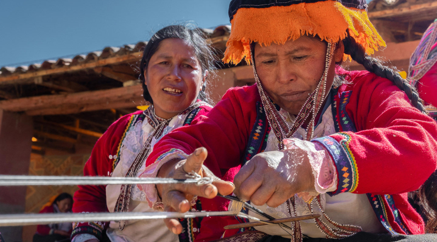 Two women hand-weaving colorful bracelets while wearing traditional Andean clothing outdoors