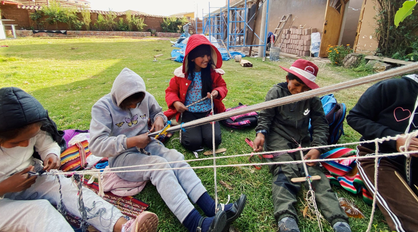 Peruvian kids weaving alpaca fiber sitting on the grass