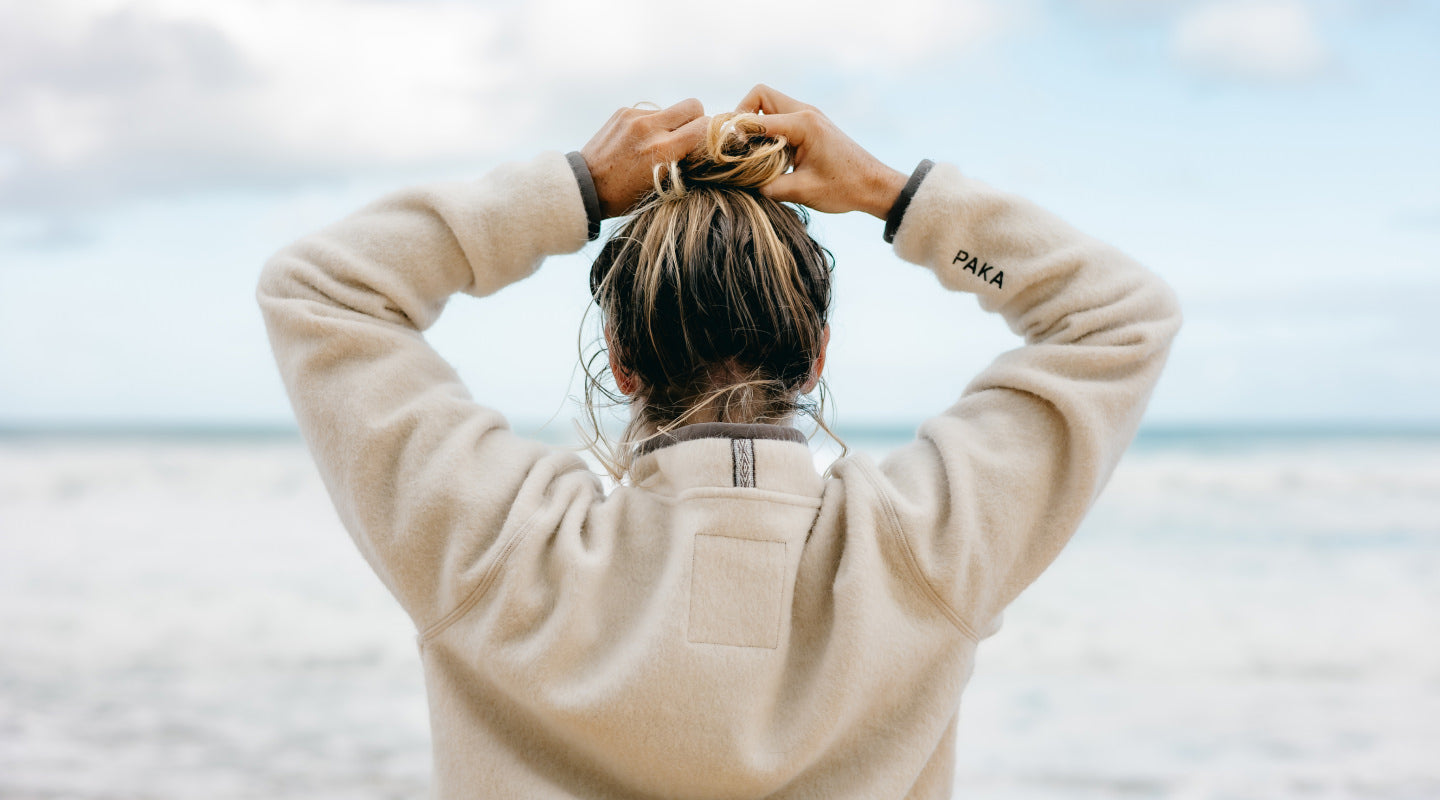 Person wearing a light tan PAKACLOUD™ alpaca fleece pullover looking out at the ocean on a windswept beach