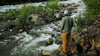 Hiker in yellow jacket standing on a rock beside a mountain stream in a green forest