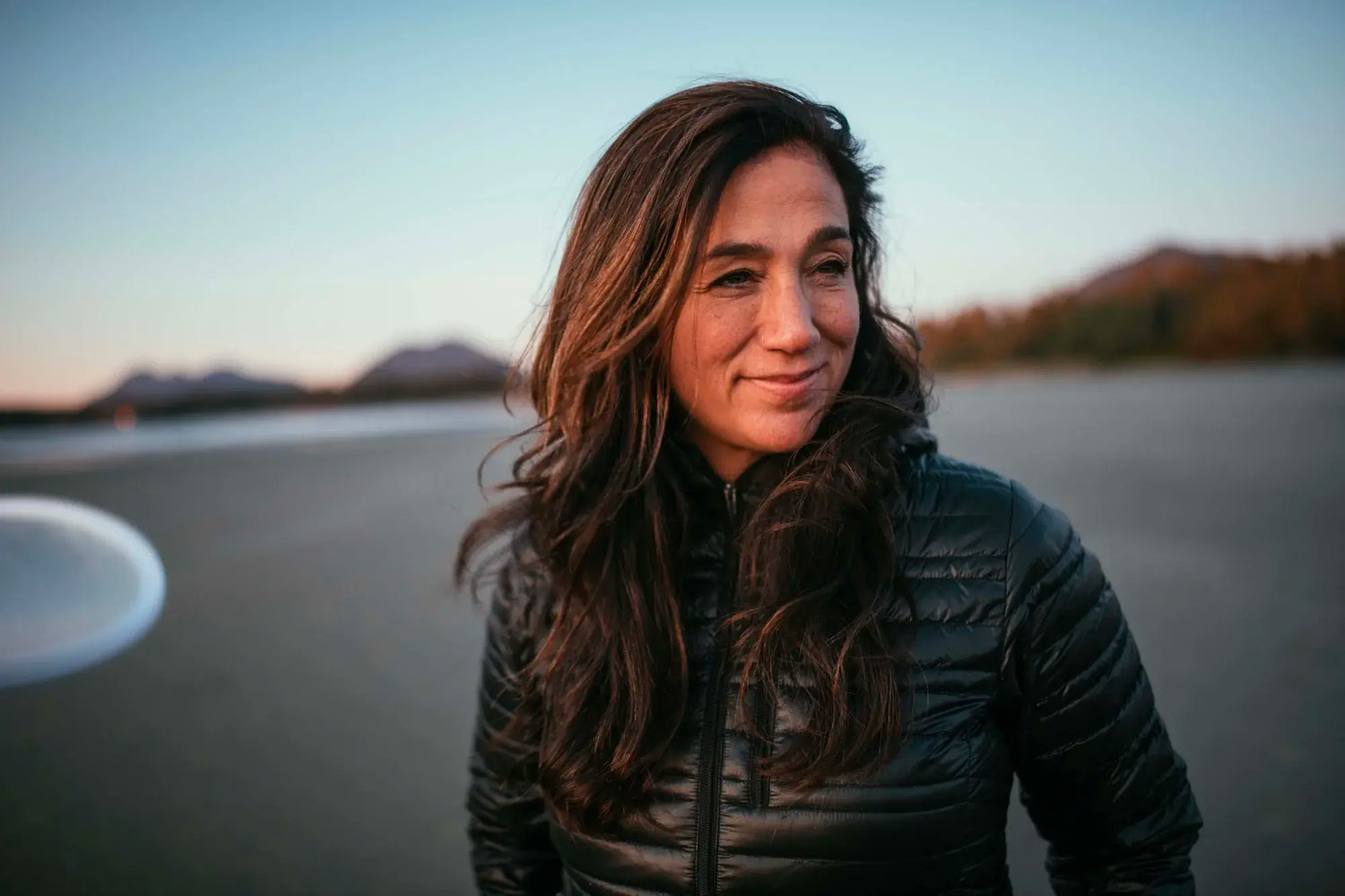 Woman standing on a beach at sunset wearing a black quilted puffy jacket, distant hills in the background