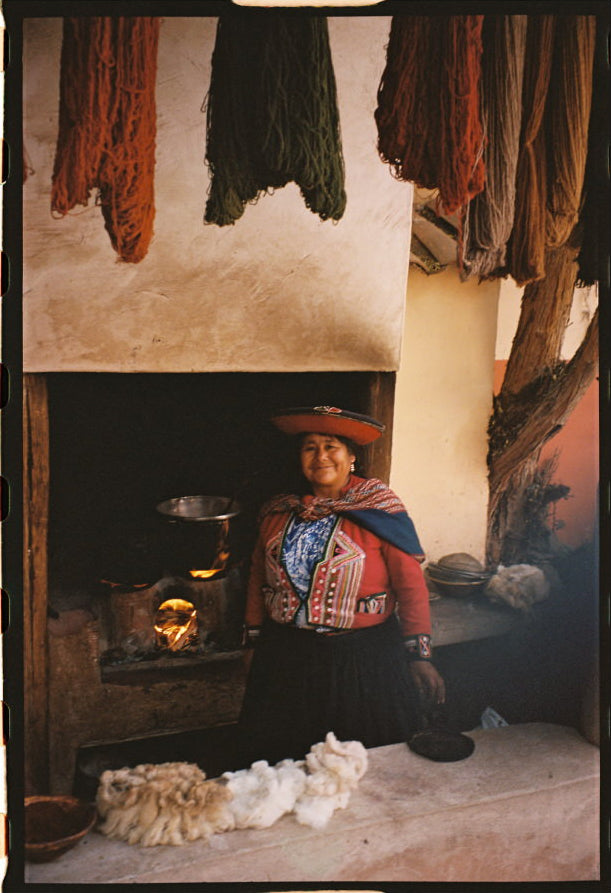 Quechua woman in traditional dress sitting by a hearth in a rustic room with hanging handwoven textiles and raw wool