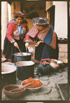 Two Quechua women stirring large dye pots in a courtyard, wearing traditional red-and-black woven garments and round hats