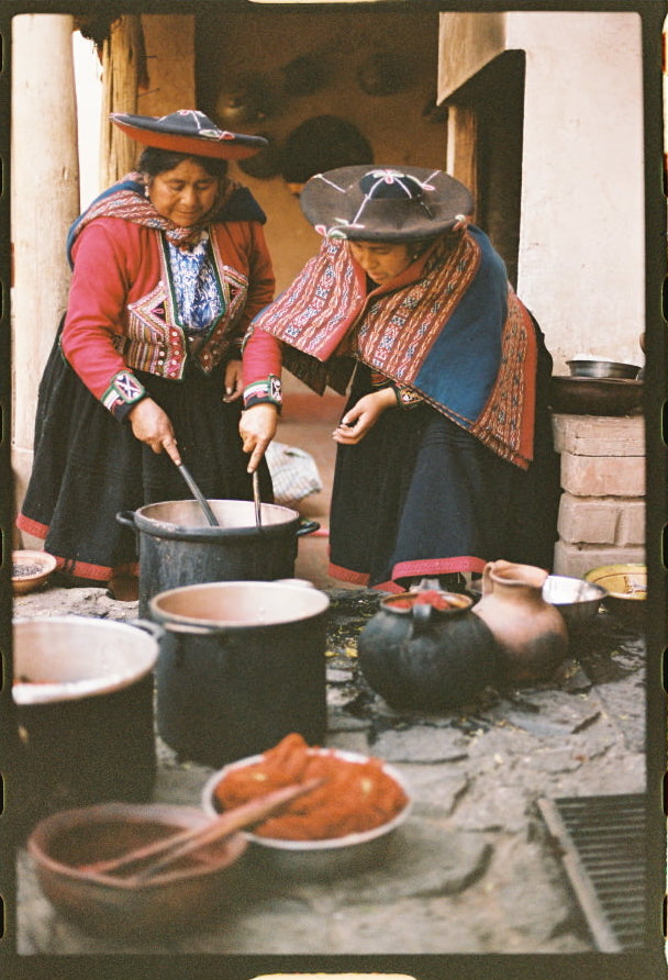 Two Quechua women stirring large dye pots in a courtyard, wearing traditional red-and-black woven garments and round hats
