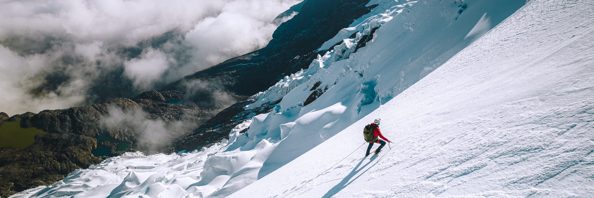Roped climber ascending a steep glacier slope in a red jacket with rocky peaks and low clouds in the background