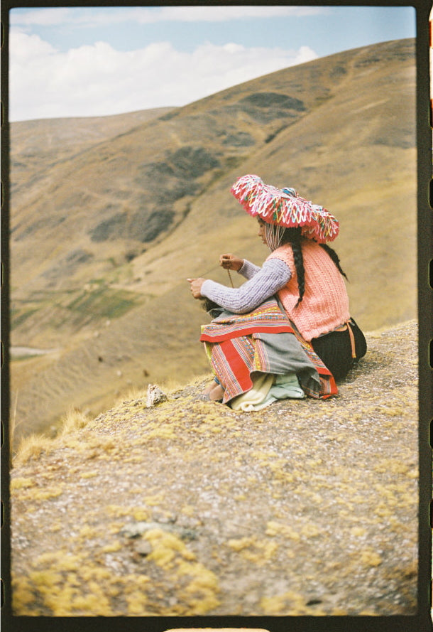 Adult wearing an orange jacket carrying a child on their back while sitting on a grassy Andean hillside