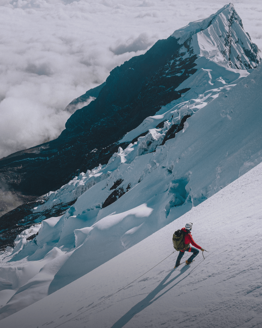 Climber descending a steep snow slope with ice axe and rope, wearing a red jacket and green pack beside glacier and clouds