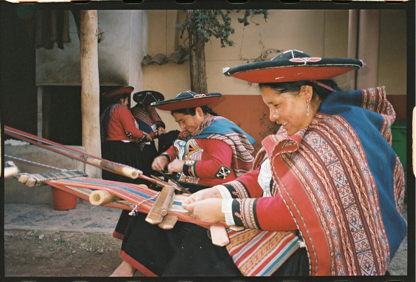 Quechua women weaving on backstrap looms in a courtyard, wearing traditional red Andean clothing and hats