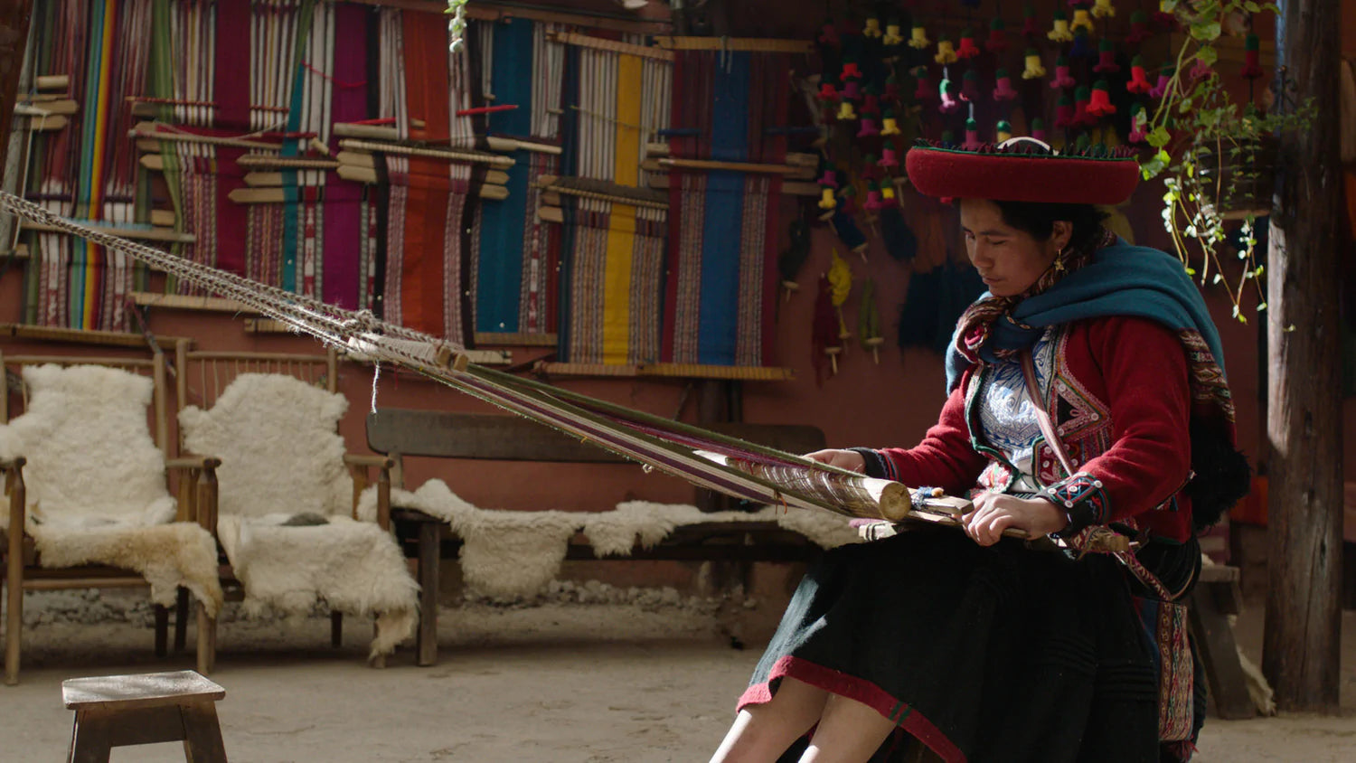 Woman weaving on a backstrap loom in a Peruvian workshop with colorful handwoven textiles hanging behind her