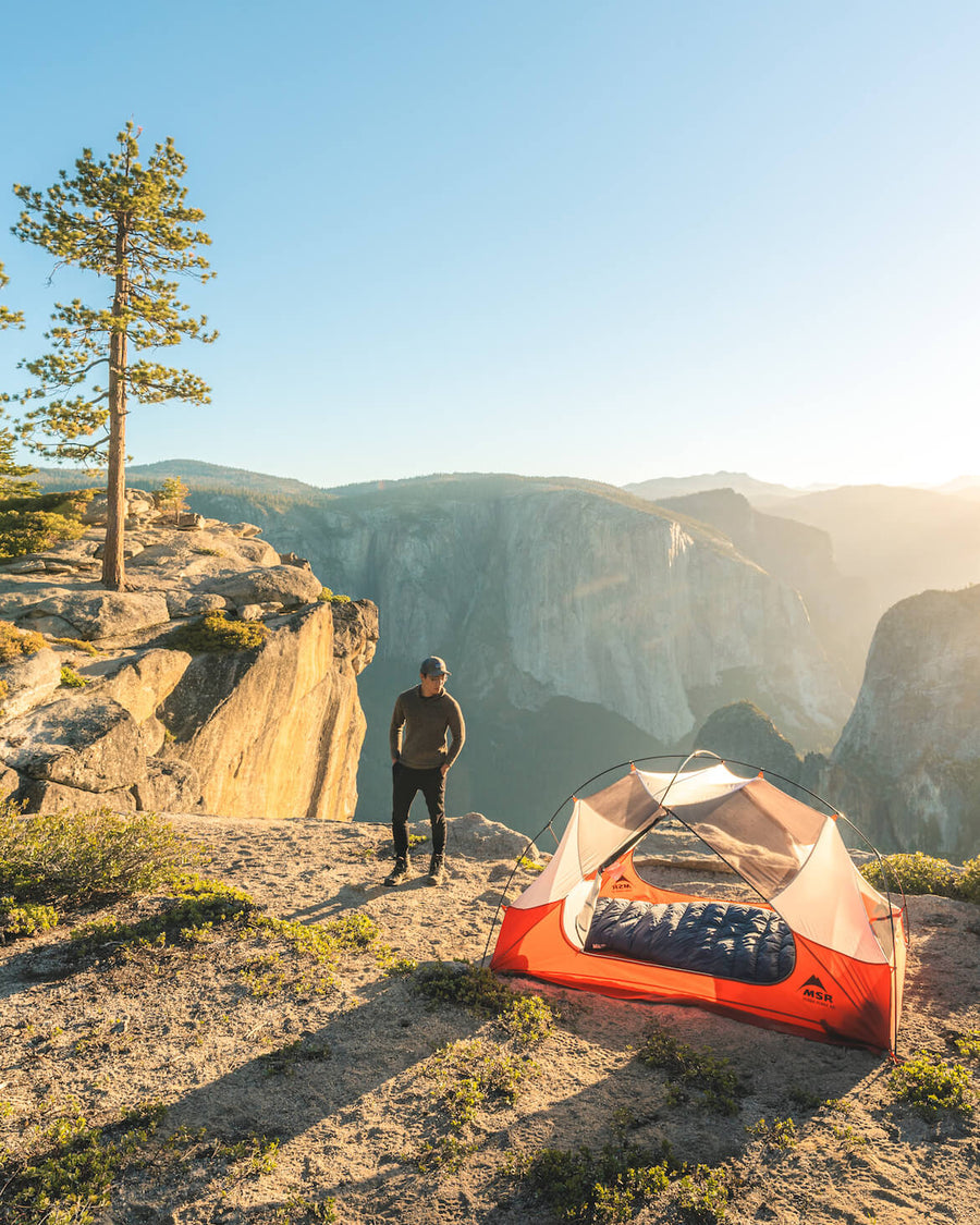 Man standing beside a tent on a cliff wearing a brown relaxed-fit The Vida hoodie with mountain valley at sunrise