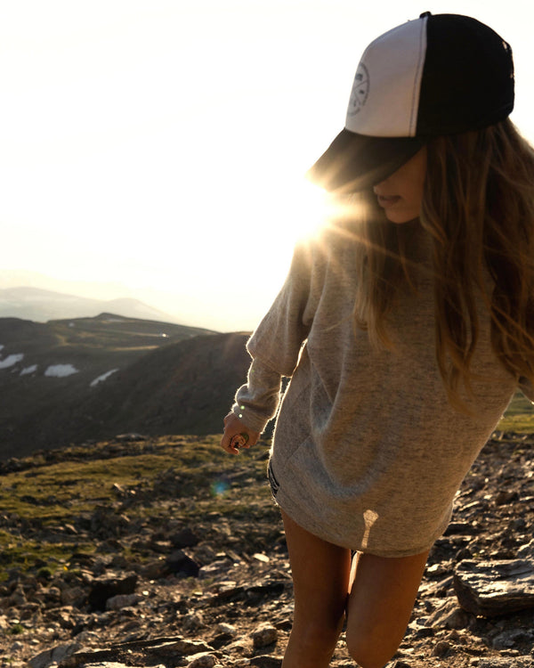Woman wearing light gray oversized alpaca hoodie and trucker cap walking on a rocky mountain trail at sunset