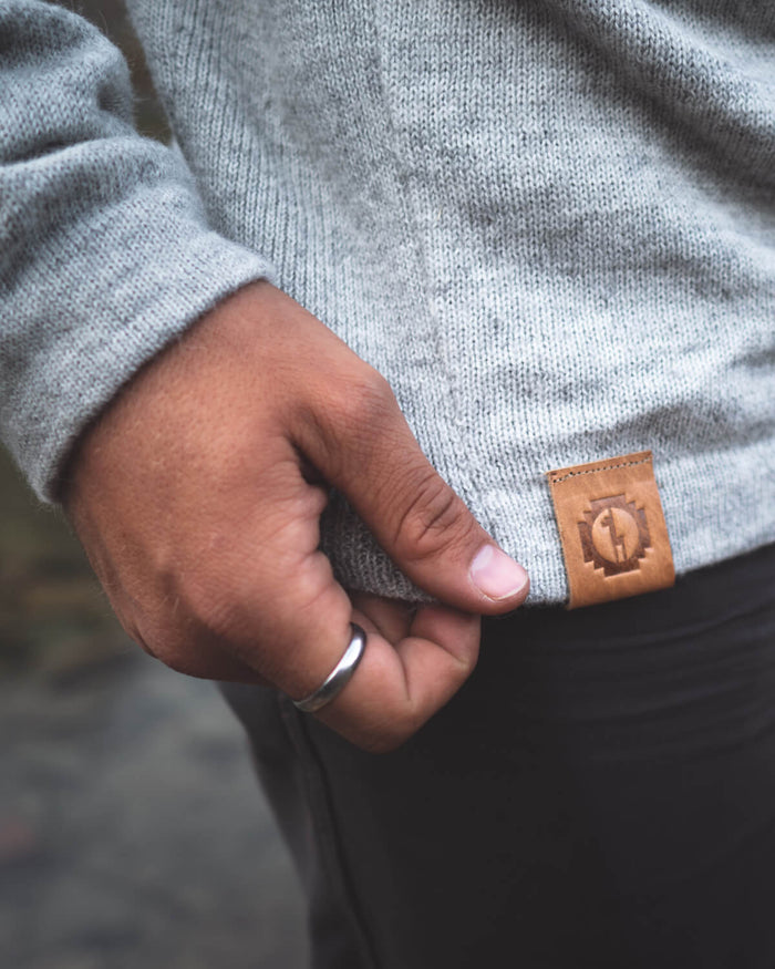 Close-up of The Hoodie in oat light tan grey showing leather logo tag at hem with hand holding the hem against dark pants