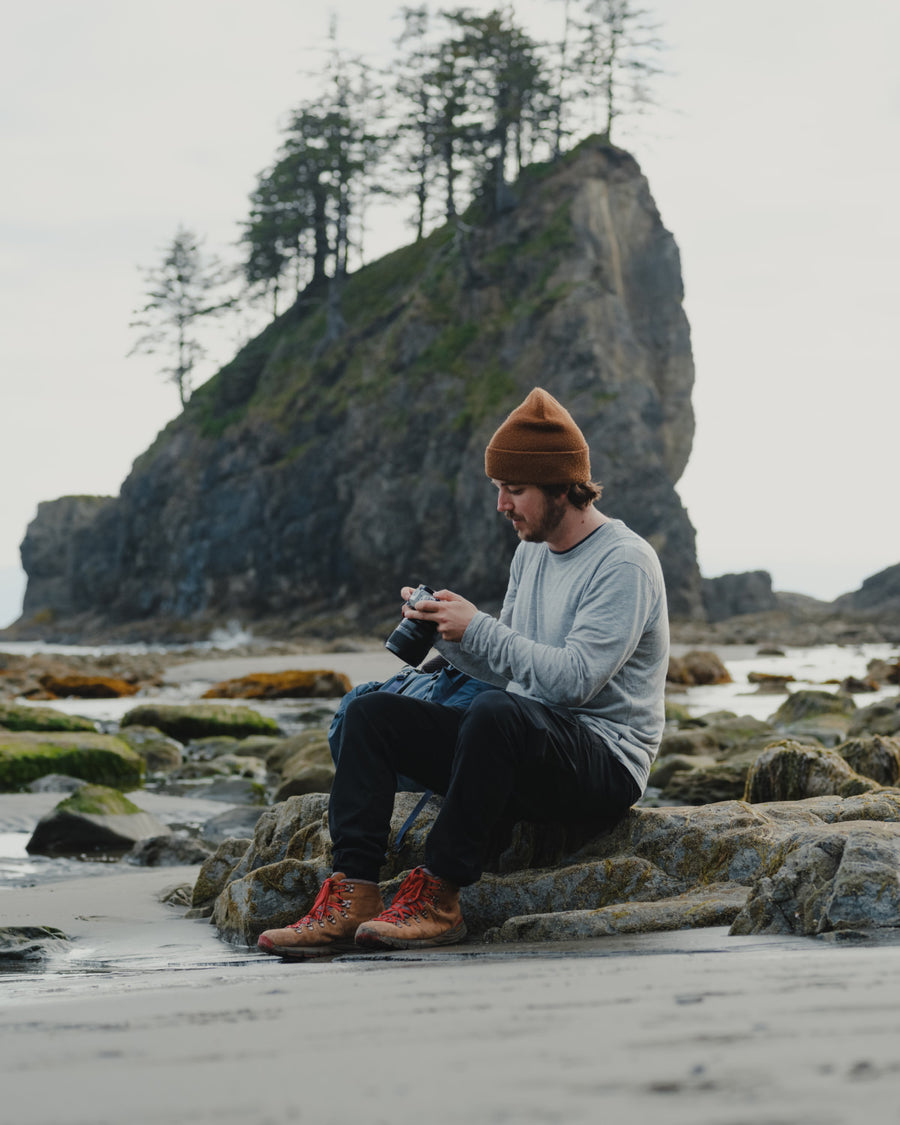 Man on Coast Looking at Camera in Front of Large Rock Formation in Heather Grey Aire Long Sleeve Shirt – PAKA Apparel