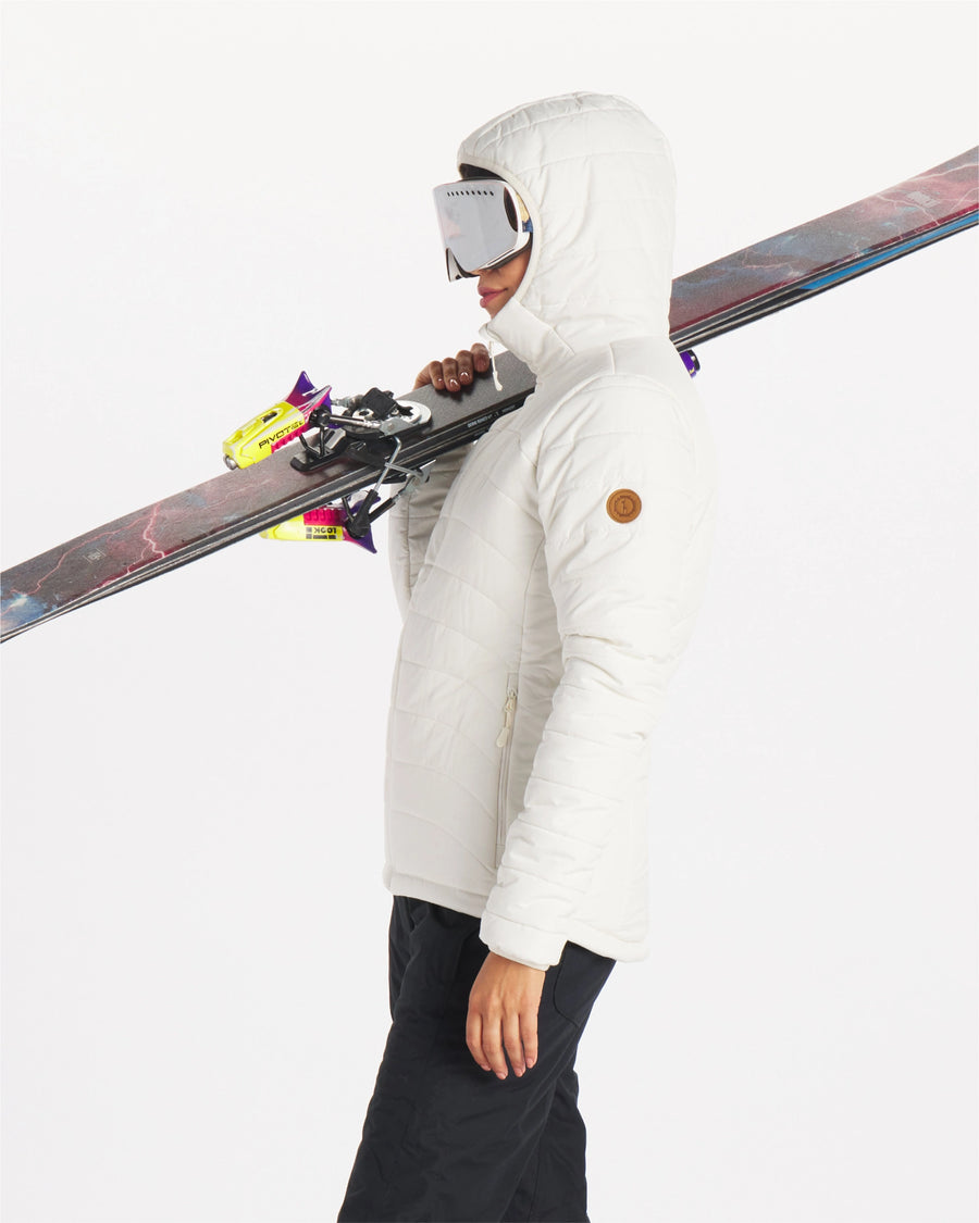 Woman wearing ivory white midweight PAKAFILL puffer jacket with hood up carrying skis over her shoulder