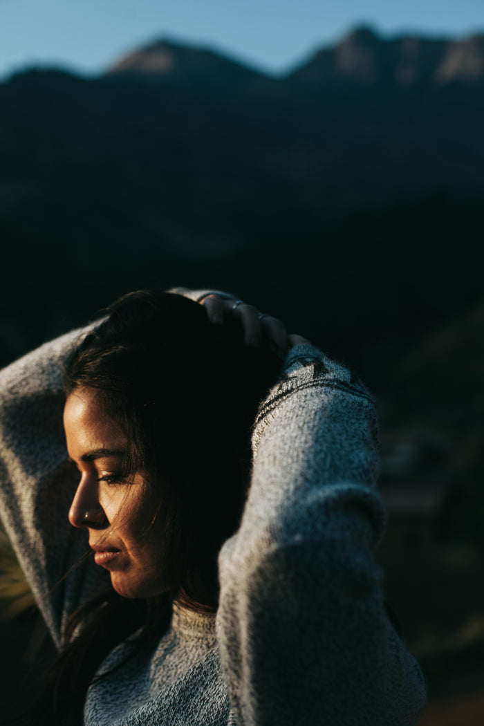 Woman wearing light grey Costa alpaca crewneck sweater in relaxed drapey fit, hands behind her head with mountains in soft evening light