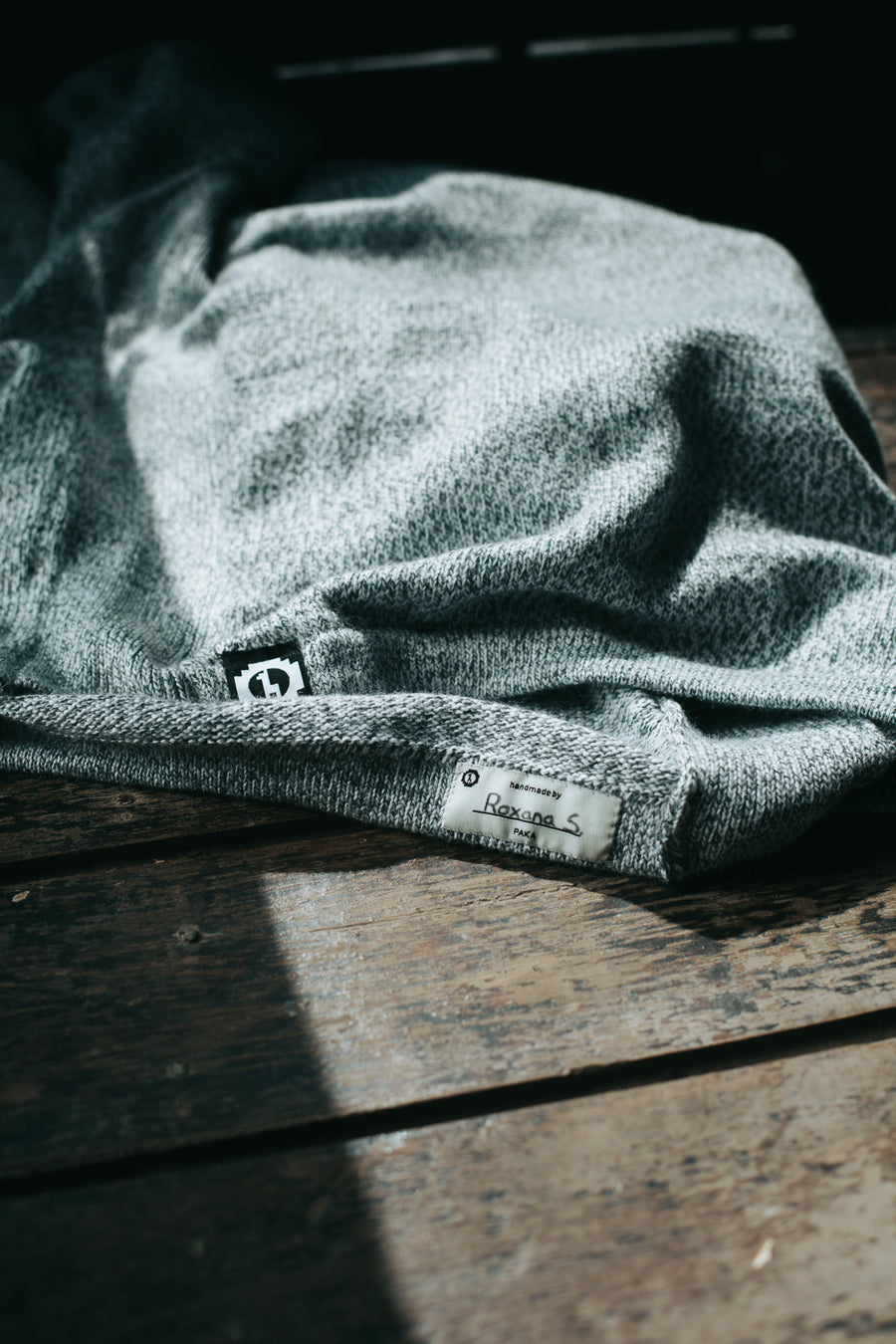Folded hem of The Costa crewneck showcasing a hand-signed artisan tag Roxana S and woven PAKA logo on a wooden surface
