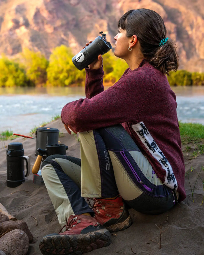 Woman sitting by a river drinking from a bottle, wearing a maroon relaxed-fit Cusco alpaca-merino sweater and hiking boots
