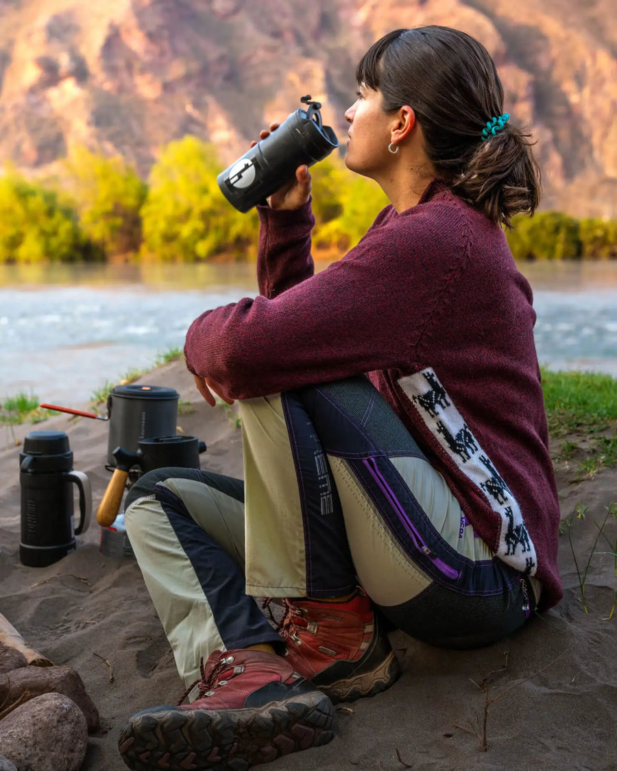 Woman sitting by a river drinking from a bottle, wearing a maroon relaxed-fit Cusco alpaca-merino sweater and hiking boots