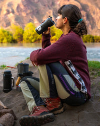 Woman sitting by a river drinking from a bottle, wearing a maroon relaxed-fit Cusco alpaca-merino sweater and hiking boots