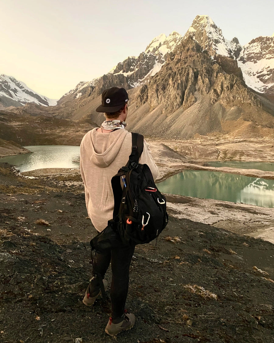 Rear view of The Hoodie in Oat showing relaxed unisex fit and hood, model with backpack by a mountain lake