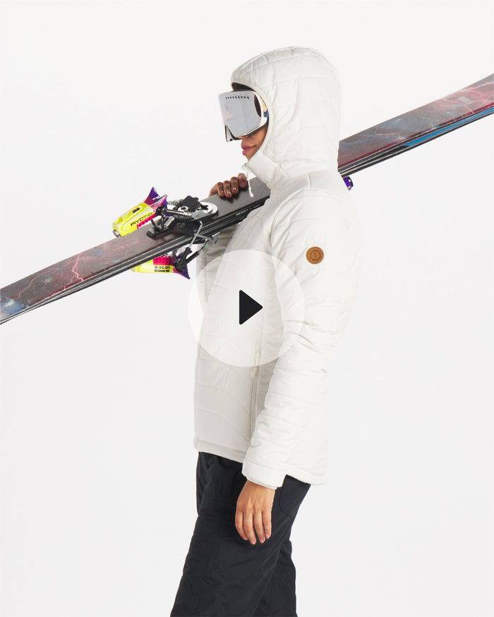 Side view of Women’s PAKAFILL Midweight Puffer with hood up, quilted alpaca insulation, model carrying skis on white background