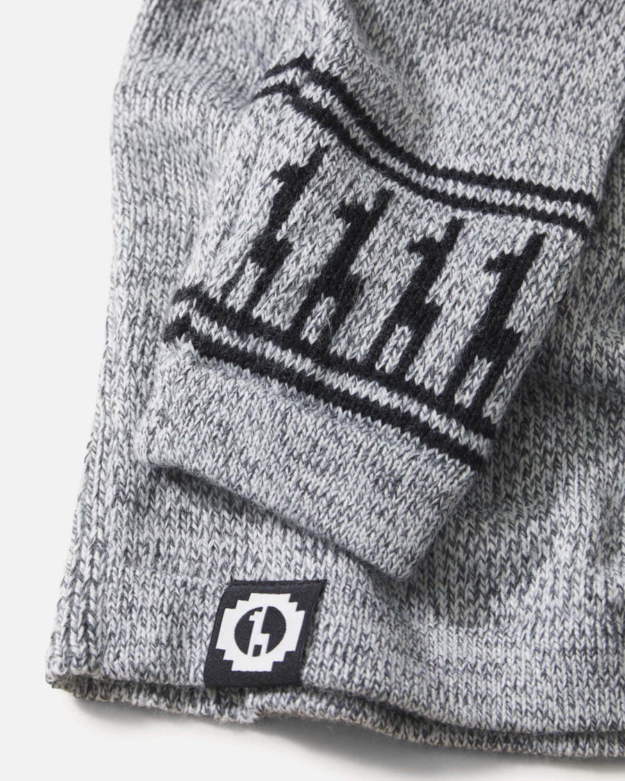 Close-up of Costa crew sweater sleeve showing marled light-grey alpaca knit, black geometric cuff pattern, and woven brand label on white background